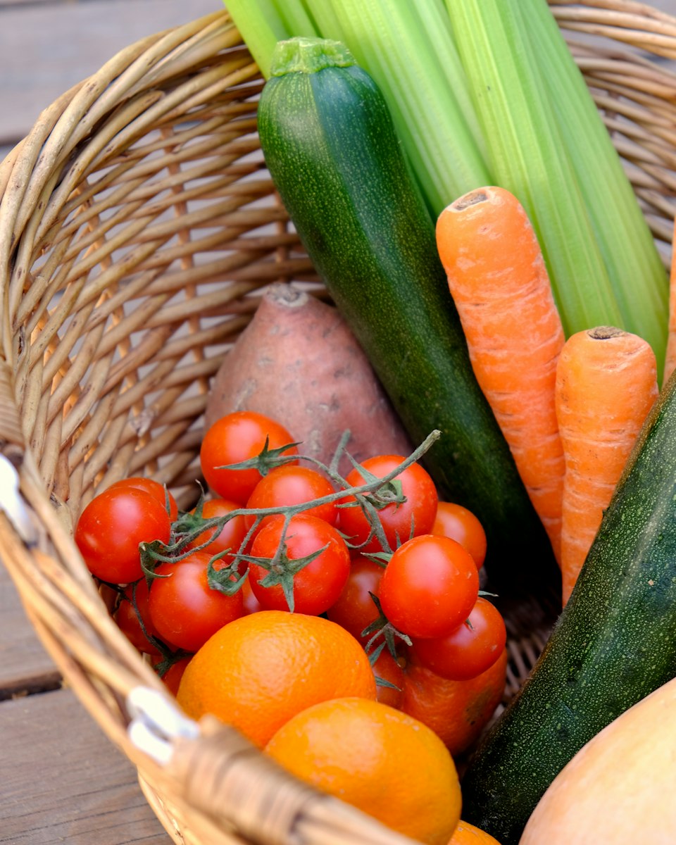 A woven basket of fresh tomatoes, carrots, zucchini, celery, oranges and a sweet potato